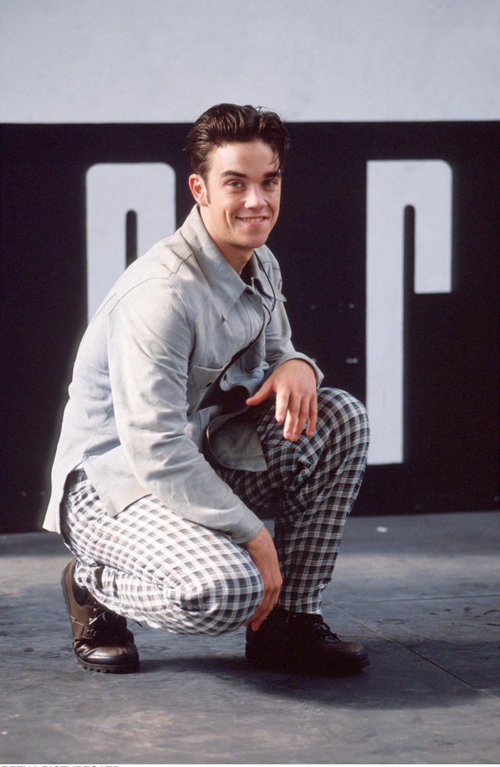 Robbie Williams crouches for a portrait in 1991, dressed in bold checkered trousers and a light jacket. Just beginning to rise as a teen idol, his playful smile and confident pose reflect the early swagger that would define his breakout years.