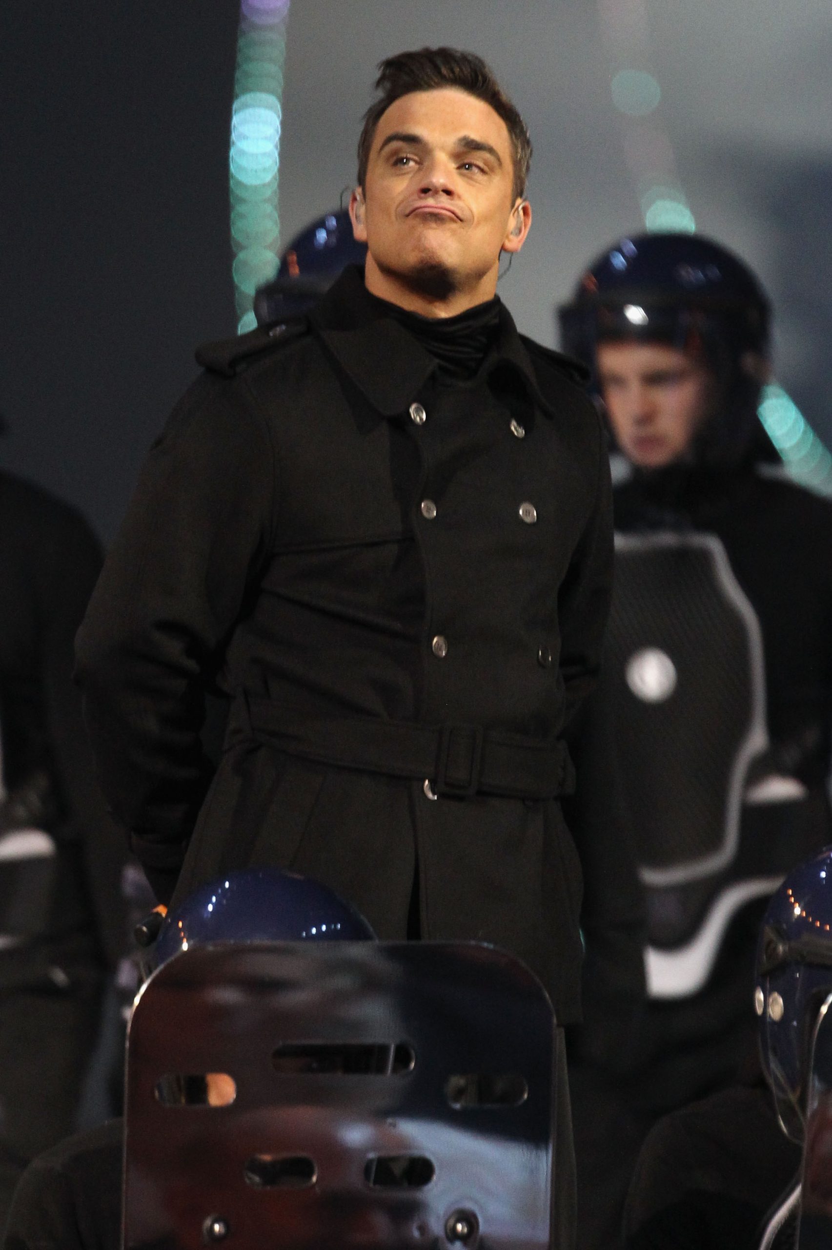Robbie Williams performed onstage at The Brit Awards 2011 on February 15 in London, England. Dressed in a belted black coat and flanked by performers in riot gear, he delivered a theatrical set that blended pop spectacle with rebellious flair.