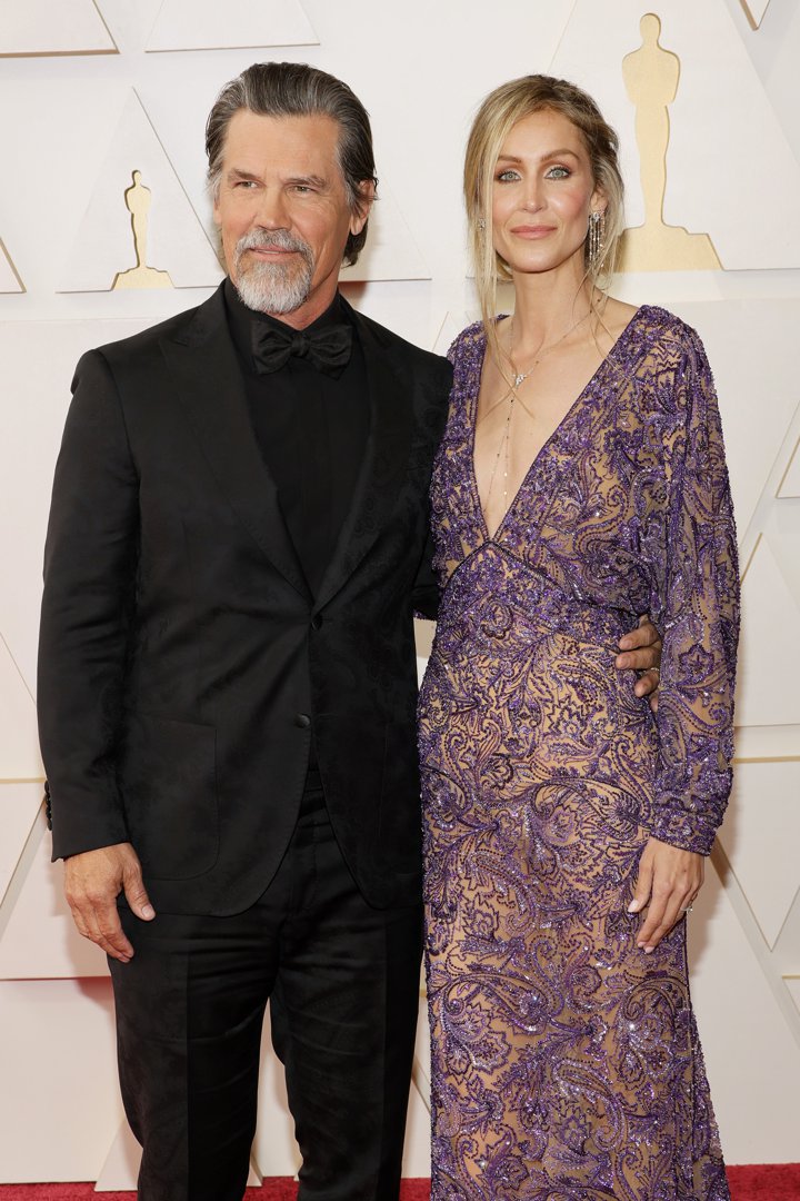 Dressed in an all-black suit with a tonal pattern and matching bow tie, he appears with a full salt-and-pepper beard and slicked-back hair. His look is mature, composed, and carefully styled.