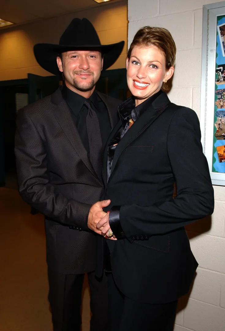 Backstage at Andre Agassi's Grand Slam fundraiser in Las Vegas, Tim McGraw and Faith Hill stand hand-in-hand, both in tailored black. Their polished look is all elegance, their pose close and quietly proud.