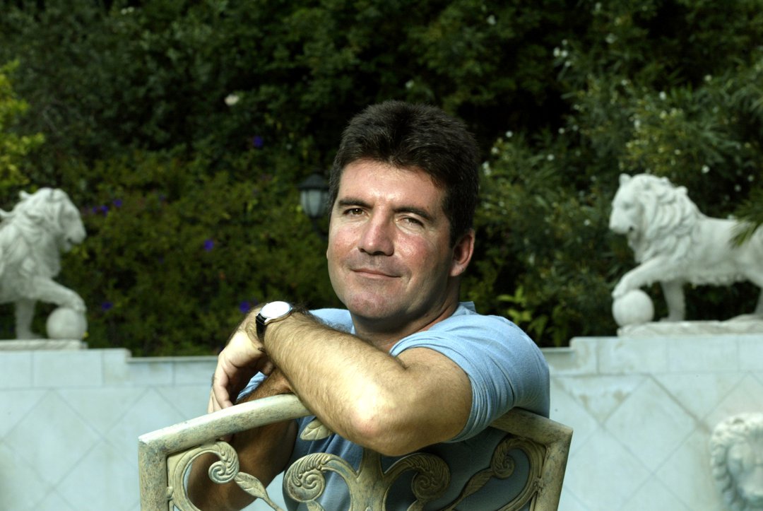 In June 2003, a rare glimpse behind the curtain &mdash; Cowell leans pensively against a garden chair at a private Beverly Hills estate. The lush backdrop and classical statues offer a stark contrast to the storm of showbiz soon to come.