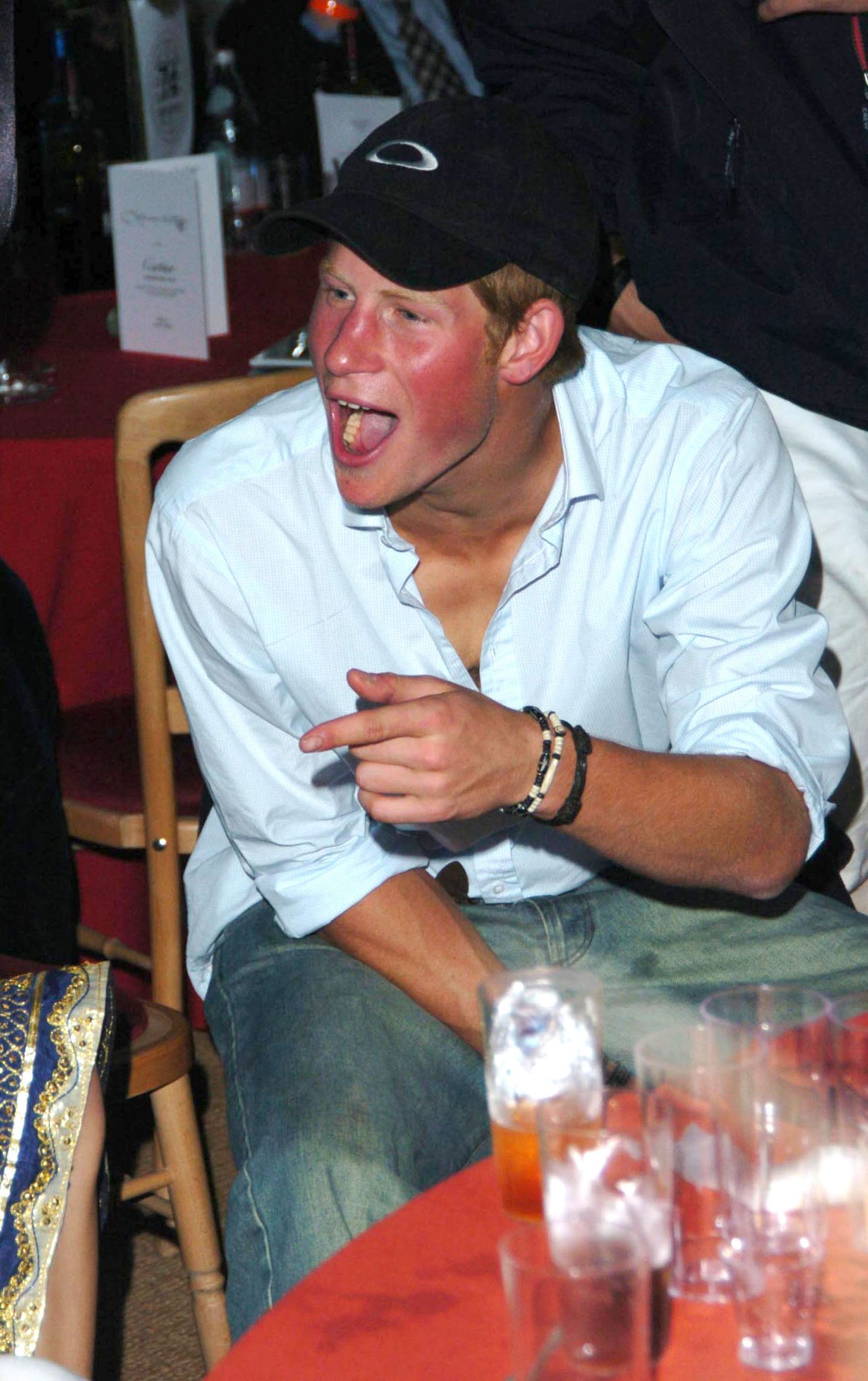 Prince Harry was photographed mid-laugh at the Guards Polo Club in Windsor on July 25, 2004, during Cartier International Day. Dressed casually in jeans, a light blue shirt, and a baseball cap, the young royal appeared entirely in his element — surrounded by friends, drinks on the table, and no shortage of mischief in the air.