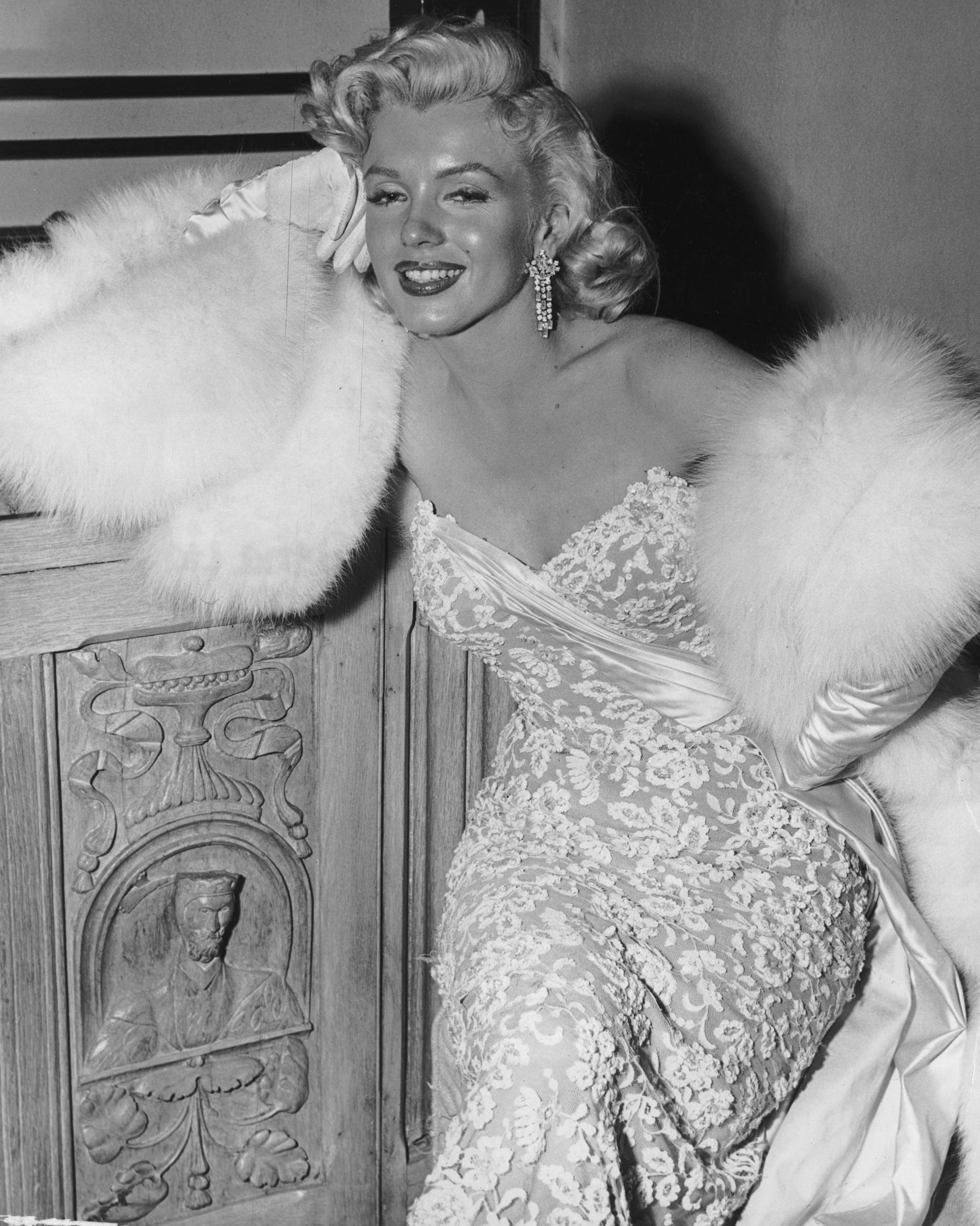 Marilyn Monroe (1926 - 1962), wearing white fur wrap and strapless evening gown, leans on a wooden cabinet during a party for the film,