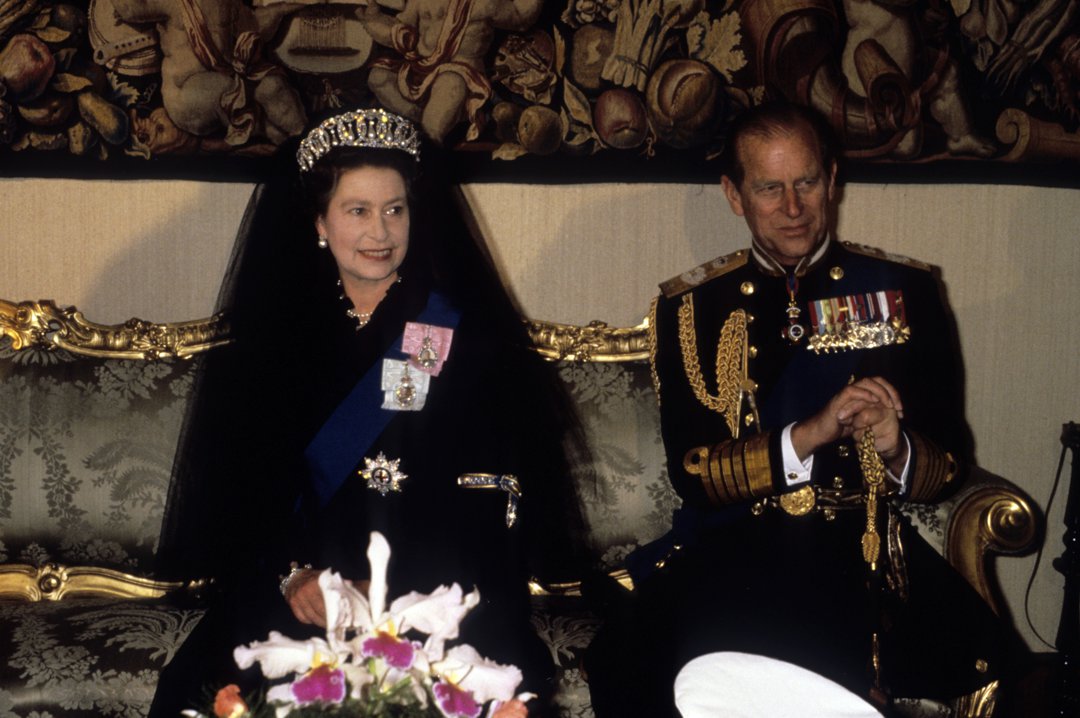 Queen Elizabeth II sits poised beside Prince Philip, Duke of Edinburgh, awaiting their historic meeting with Pope John Paul II at the Vatican on 17 October 1980. Crowned in a glittering tiara and royal regalia, the monarch embodies grace and dignity, while the Duke appears in full military dress.