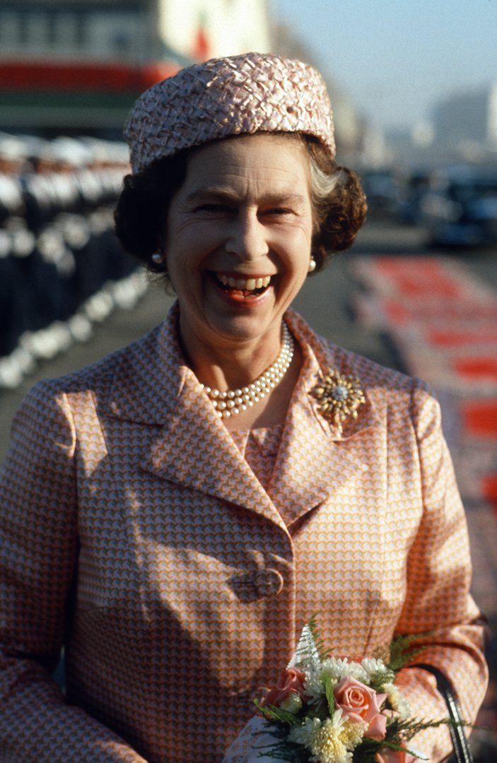 Queen Elizabeth II shares a rare moment of unguarded laughter as she arrives in Casablanca on 27 October 1980. With a floral corsage pinned to her elegant patterned coat and a pillbox hat perched perfectly, the Queen radiates warmth on this unforgettable royal tour.