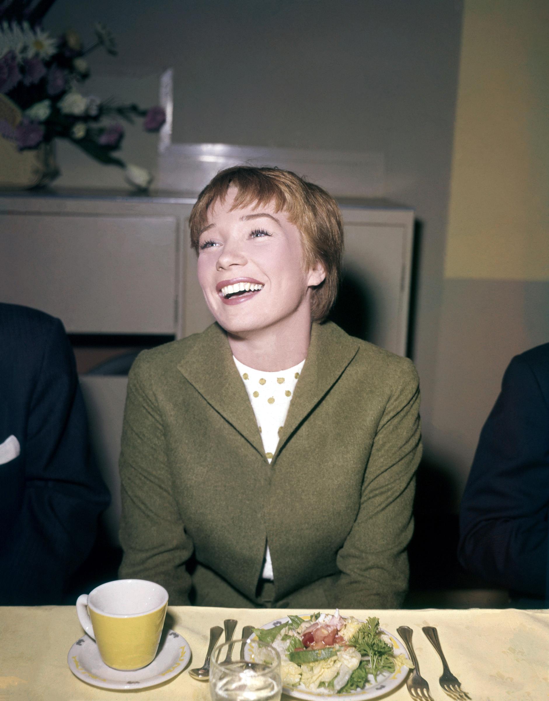 A young Shirley MacLaine lights up the room during a lunch in Los Angeles, circa 1959. Draped in a smart green jacket and patterned blouse, the rising star is caught mid-laughter &mdash; her charm and charisma already undeniable as she soared to Hollywood stardom.