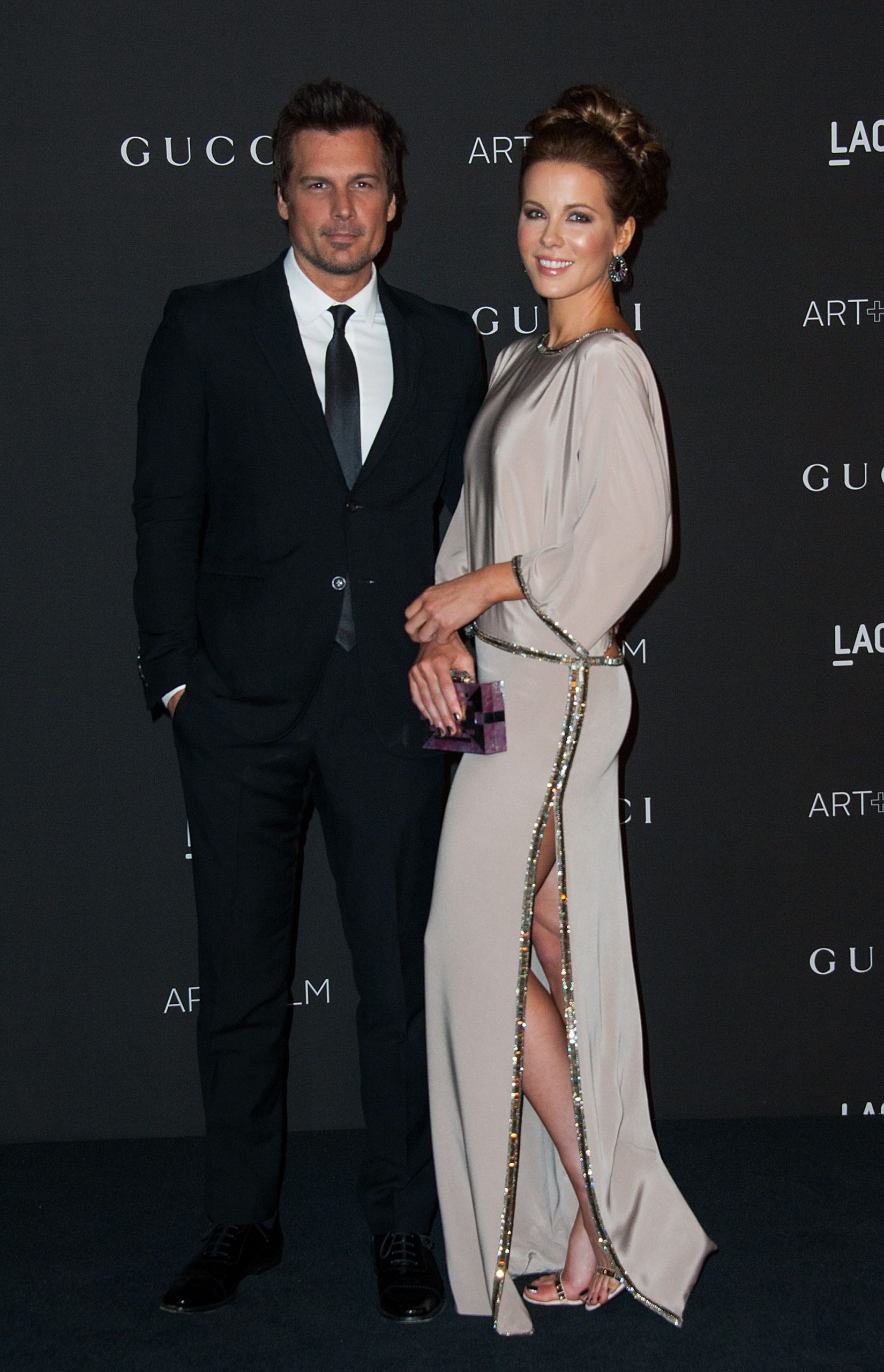 Kate Beckinsale poses alongside her then-partner at a Gucci-sponsored event, wearing a flowing pale gown with a thigh-high slit and embellished trim, her hair styled in an elegant updo and accessorised with statement earrings.