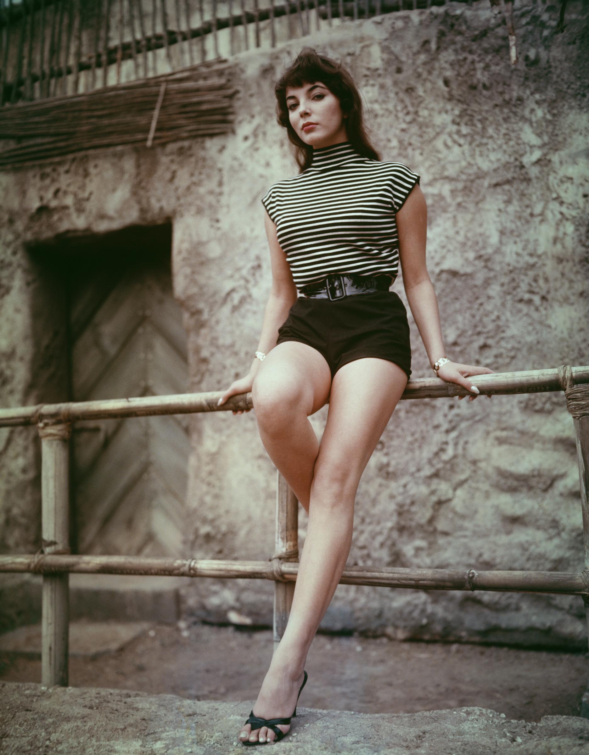 Poised with early-career confidence, Joan Collins exuded classic mid-century glamour in this circa 1955 portrait. Wearing high-waisted shorts and a striped sleeveless top, she leaned effortlessly against a bamboo railing, her heels and polished posture capturing the youthful allure that helped launch her into international stardom.