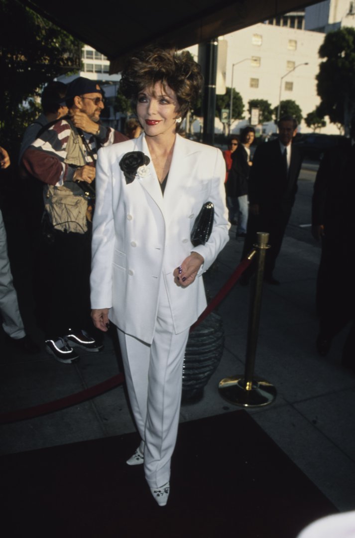 Evergreen Creator said:  Joan Collins arrived at the Beverly Hilton Hotel on June 7, 1999, for the R.A.D.D. Benefit Gala honouring Liza Minnelli, dressed in a crisp white double-breasted suit adorned with a dramatic black flower brooch. Her structured tailoring, glossy clutch, and signature voluminous hair captured a refined take on late-1990s red carpet elegance.