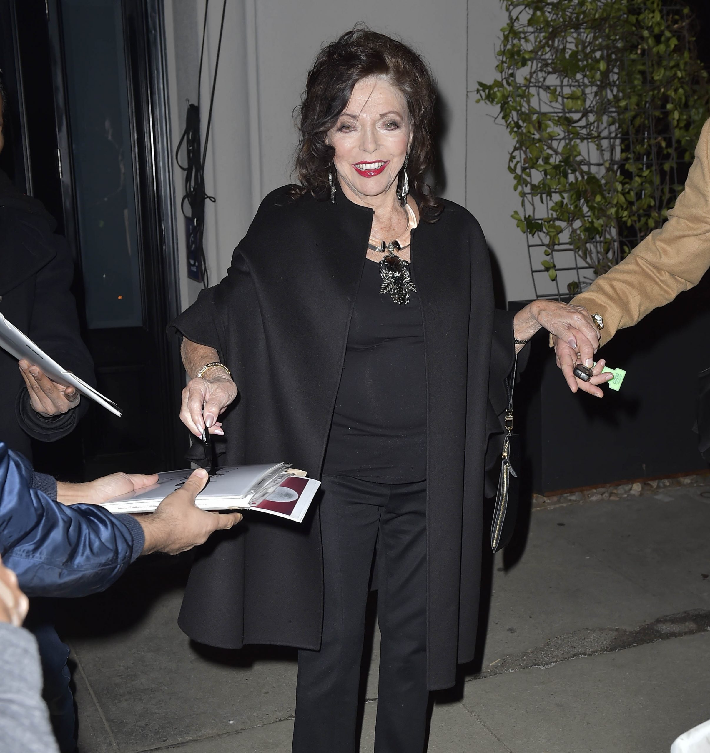 Joan Collins was seen in Los Angeles, California, on February 8, 2022, dressed in an all-black ensemble that balanced elegance with ease. Draped in a structured cape and accessorised with a statement brooch and drop earrings, she greeted fans warmly, effortlessly commanding attention with her enduring presence and polished poise.