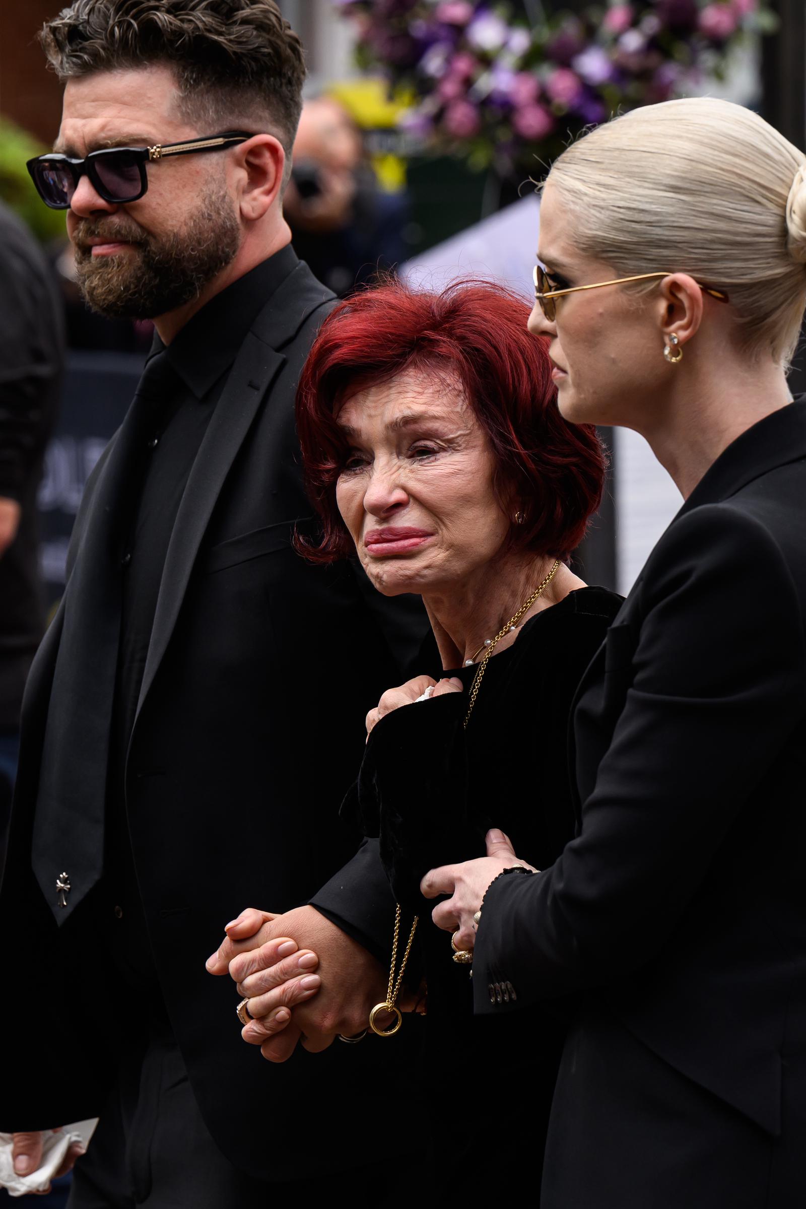 Sharon Osbourne is supported by her children Jack and Kelly Osbourne as she attends a memorial event, her expression marked by visible emotion.