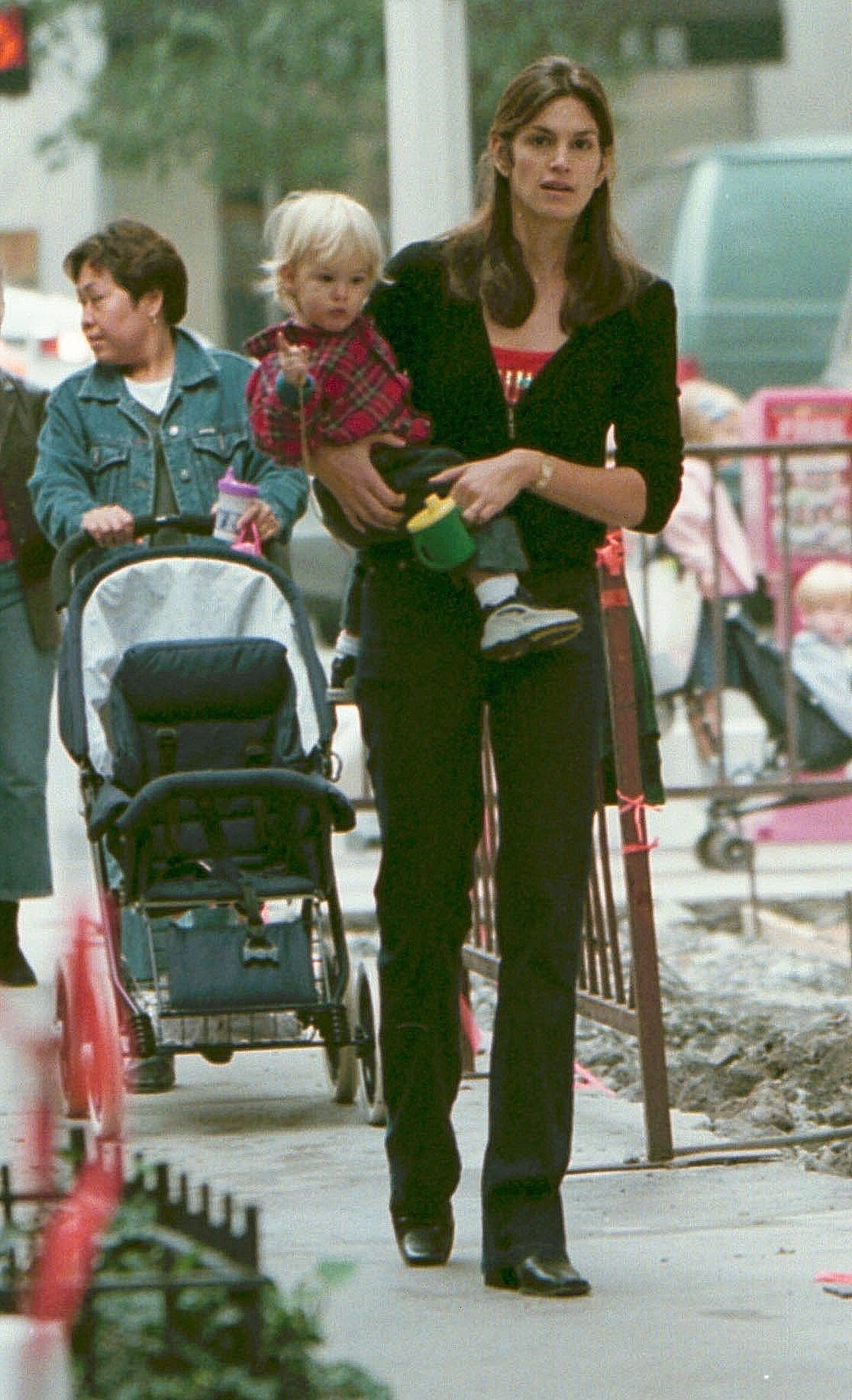 Cindy Crawford carries a toddler Presley Gerber through midtown Manhattan on October 27, 2000, during a casual city stroll. With his white-blonde hair, tiny trainers, and flannel shirt, Presley clutches a green sippy cup as Crawford walks confidently by his side — a candid glimpse of early motherhood set against the backdrop of busy New York life.