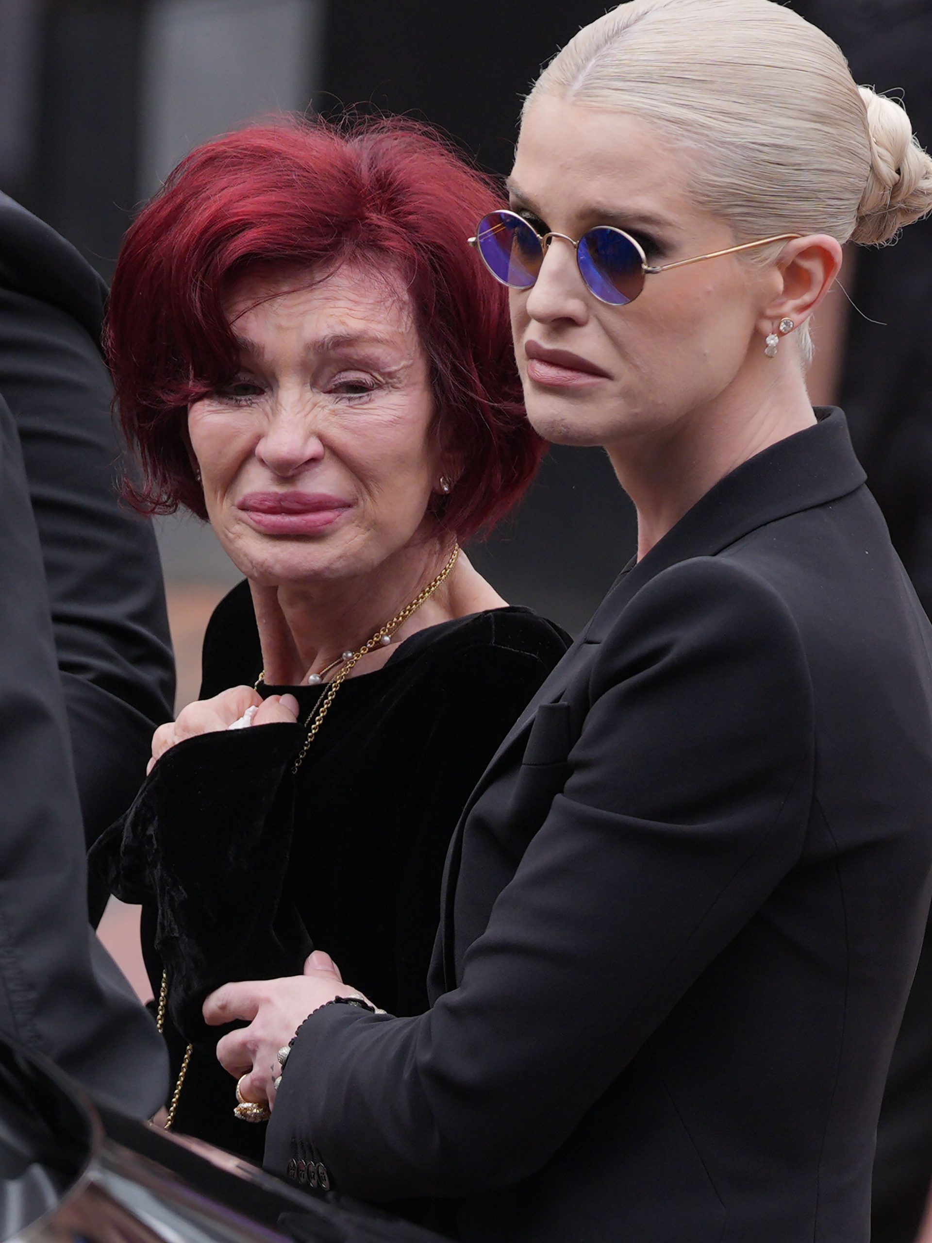 Sharon Osbourne and Kelly Osbourne stop to view tributes to the late Ozzy Osbourne from fans at Black Sabbath Bench and Bridge as his funeral cortege travels through his home city of Birmingham on 30 July 2025 in Birmingham, England. | Source: Getty Images