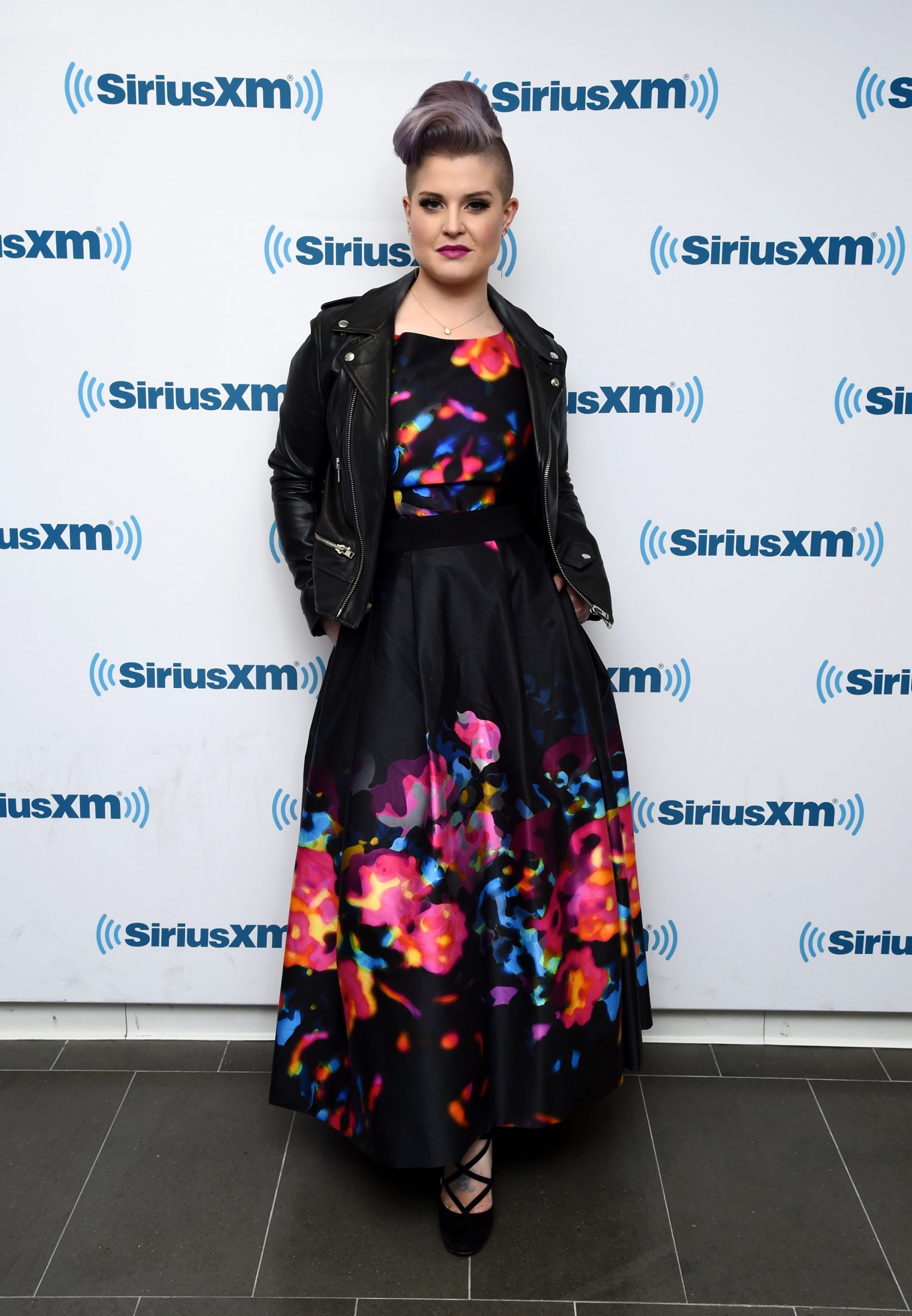 Turning heads at SiriusXM Studios on 12 November 2015, Kelly blends punk edge with high fashion, rocking her signature lavender mohawk, a fierce leather jacket, and a vibrant, abstract-print dress — unapologetically bold.