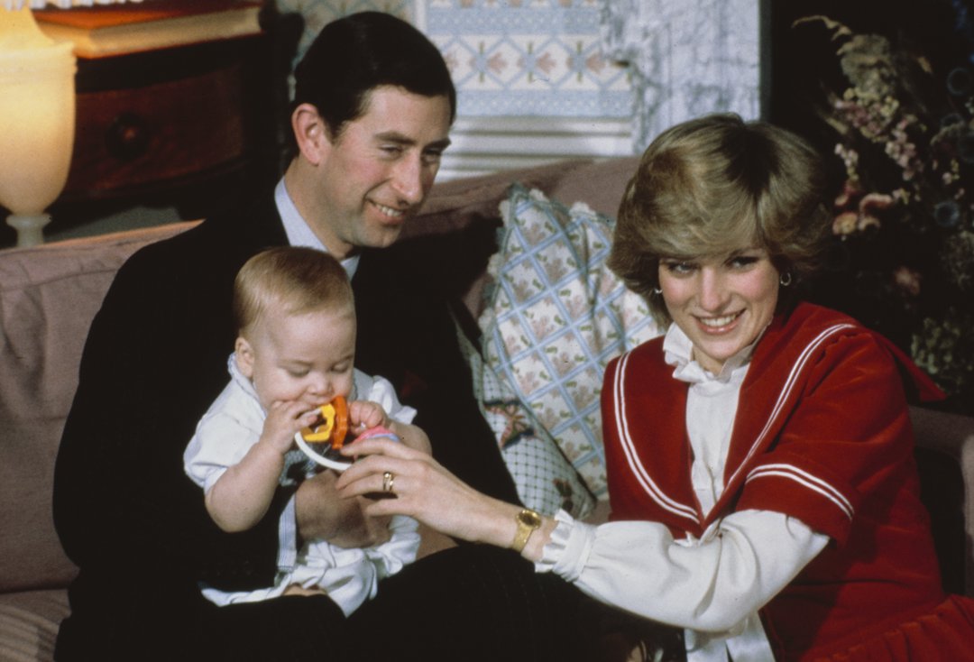 Captured at Kensington Palace on December 22, 1982, this tender family portrait shows King Charles III, then the Prince of Wales, cradling a young Prince William as Diana, former Princess of Wales, gently offers a teething ring. Dressed in a red-and-white sailor-style outfit, the Princess radiates youthful warmth and joy.