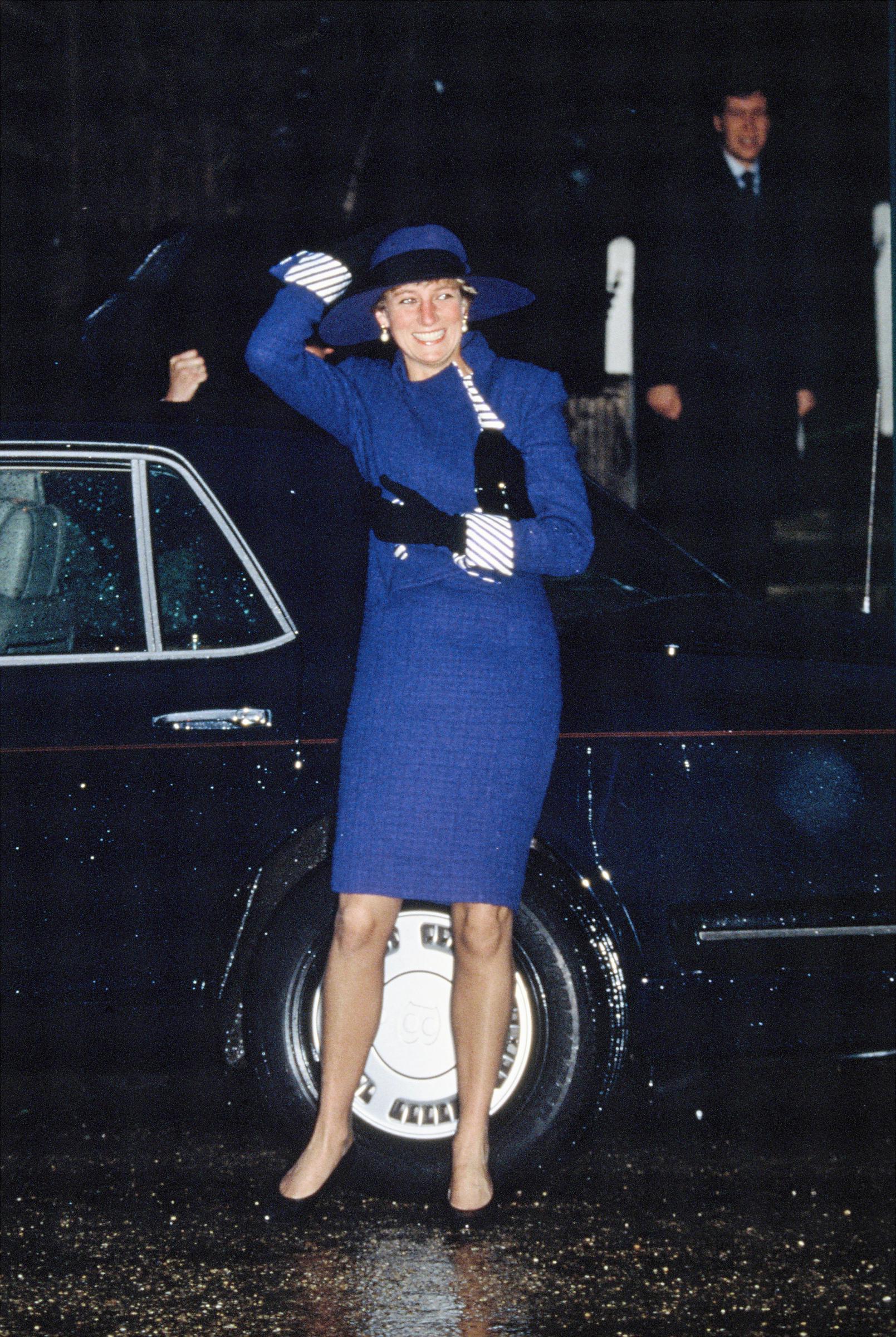 On a rainy Christmas morning in Sandringham, the Princess is seen arriving at church for the traditional royal service on December 25, 1990. Dressed in a striking cobalt ensemble with matching wide-brimmed hat and striped accessories, she shields herself from the wind with a smile that outshone the weather.