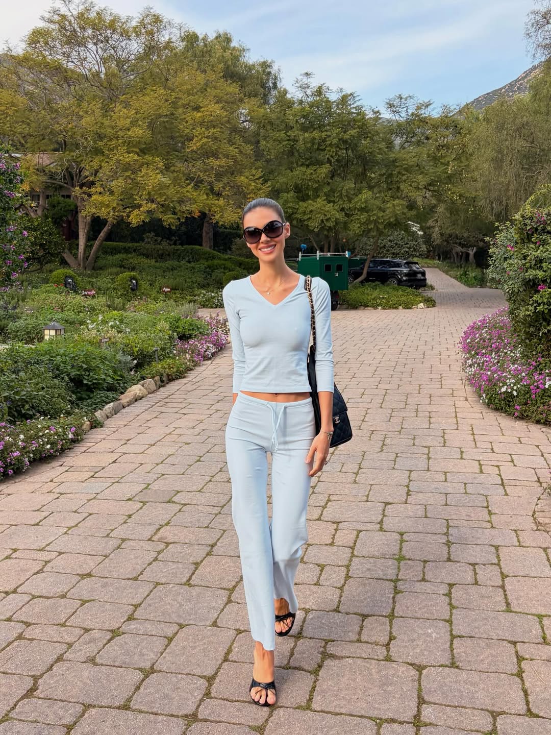 Nicola Peltz Beckham keeps it effortlessly chic in a tranquil garden stroll, wearing a soft blue long-sleeved top and matching trousers. Dark sunglasses and a sleek shoulder bag complete the look, while the minimalist styling lets the serene surroundings take center stage. | Source: Instagram/nicolapeltzbeckham