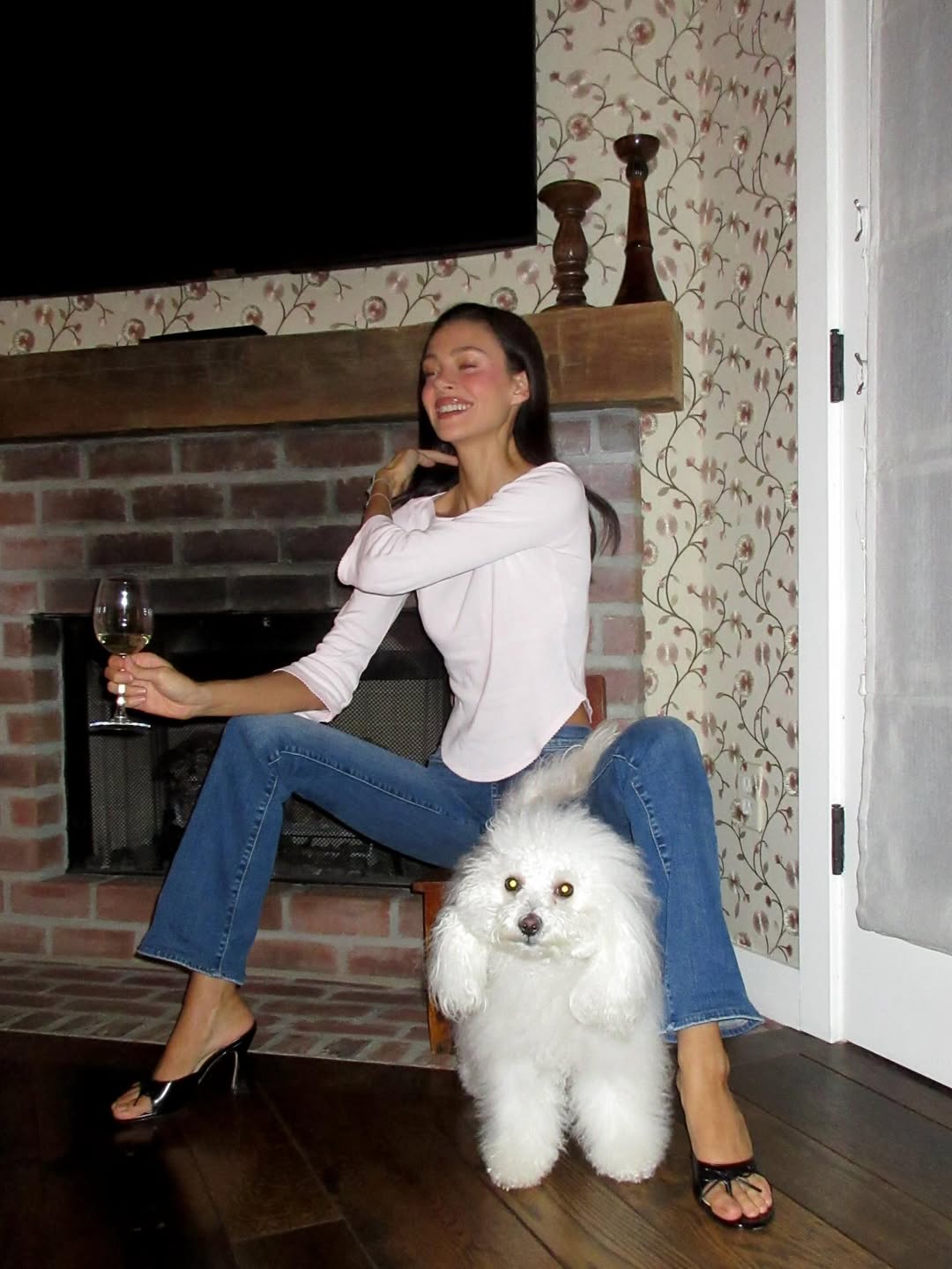 At home by the fireplace, Nicola shares a cozy evening with a glass of wine and her dog curled up at her feet. Flipping her hair back with elegance, she laughs for the camera in a relaxed, candid moment. | Source: Instagram/nicolapeltzbeckham