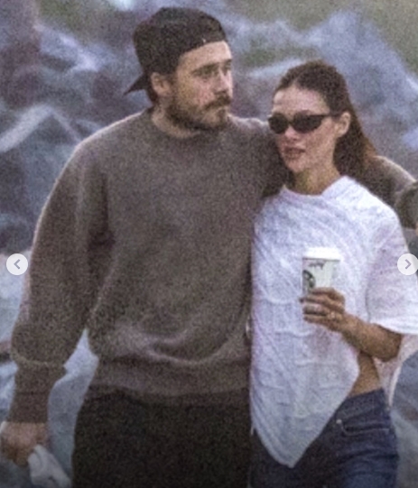 Brooklyn Peltz Beckham wraps an arm around Nicola Peltz Beckham as the couple walks along the beach in Malibu. Nicola holds a Starbucks cup and wears sunglasses, while Brooklyn keeps close in a brown sweater and backwards cap. | Source: Instagram/dailymailuk