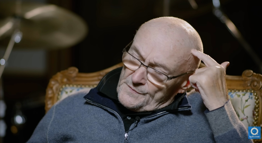 Wearing a zip-up jumper and seated in an ornate chair, Collins lowers his eyes and gestures toward his head — a subtle but telling glimpse into the physical and emotional wear that comes with years of chronic illness. Despite a long list of health challenges, he maintains a quiet dignity in his presence. | Source: YouTube/Drumeo