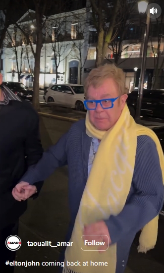 Wearing bright blue glasses and a yellow scarf, Sir Elton John appears tense as he makes his way past cameras. | Source: Instagram/taoualit__amar