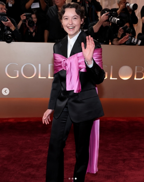 Bella Ramsey waves on the red carpet while attending the 83rd annual Golden Globe Awards wearing a tailored black suit accented by a striking pink sash and trailing fabric detail, from a post dated January 12, 2026. | Source: Instagram/justjared