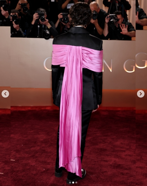 A rear view shows Bella Ramsey's dramatic pink sash flowing down the back of their black suit as they pose on the red carpet at the 83rd annual Golden Globe Awards in Beverly Hills, California, from a post dated January 12, 2026. | Source: Instagram/justjared