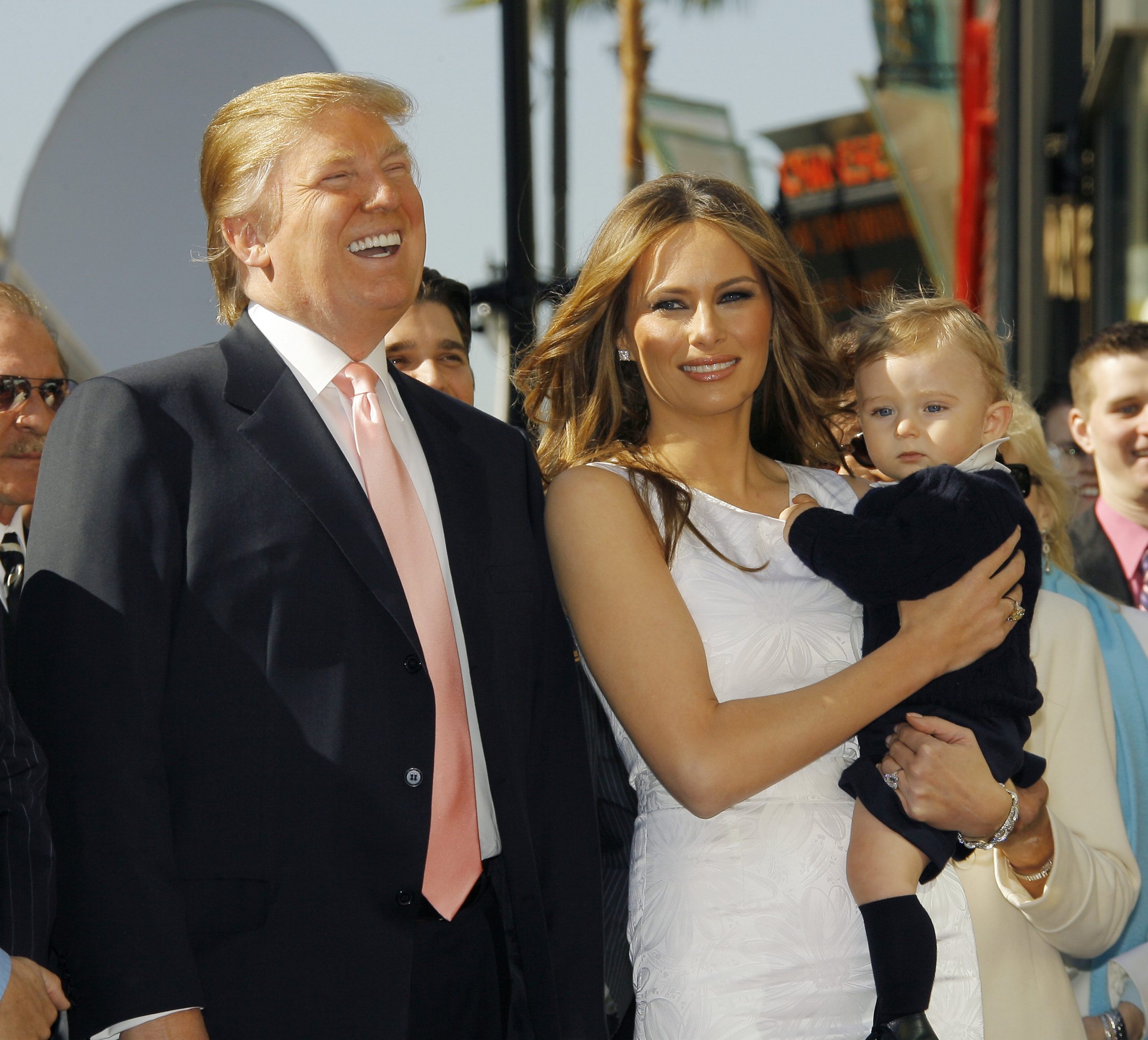 At his Hollywood Walk of Fame ceremony, Melania Trump holds baby Barron in her arms as cameras flash. Standing beside them, Donald Trump smiles toward the gathered crowd. The family poses together during the moment he is honored with his star.