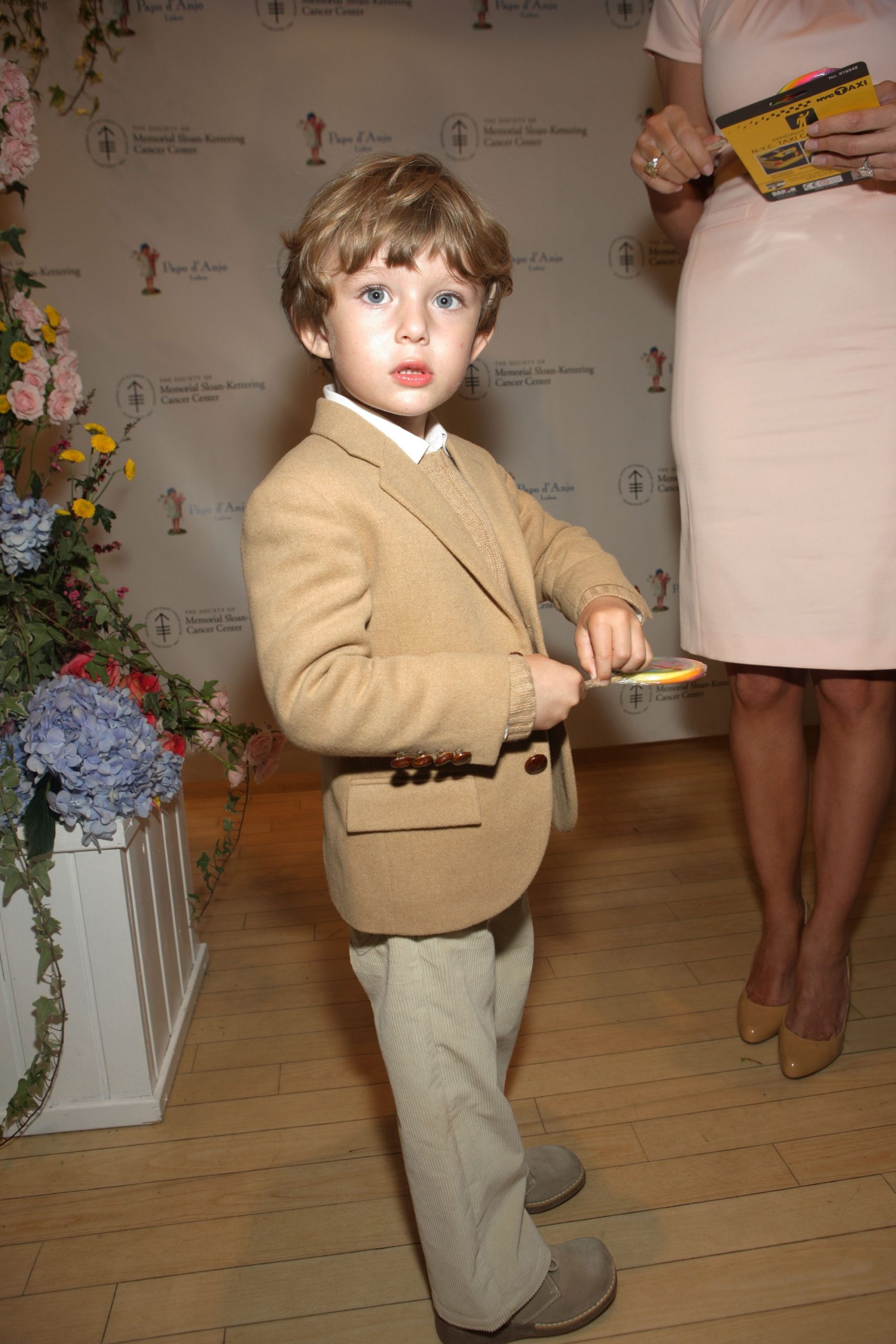 At the 18th Annual Bunny Hop, Barron Trump steps forward in a tan blazer layered over a knit sweater and collared shirt. He pairs the look with light corduroy trousers and soft gray shoes, standing out against the floral display nearby.