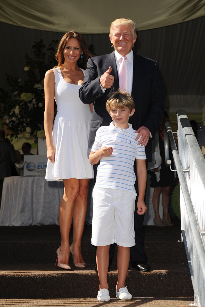At the Trump Invitational Grand Prix at Mar-a-Lago, Donald Trump stands beside Melania Trump and their son, Barron Trump, on the venue steps. Melania wears a sleeveless white dress and nude heels, while Barron appears in a striped polo shirt with white shorts and loafers.