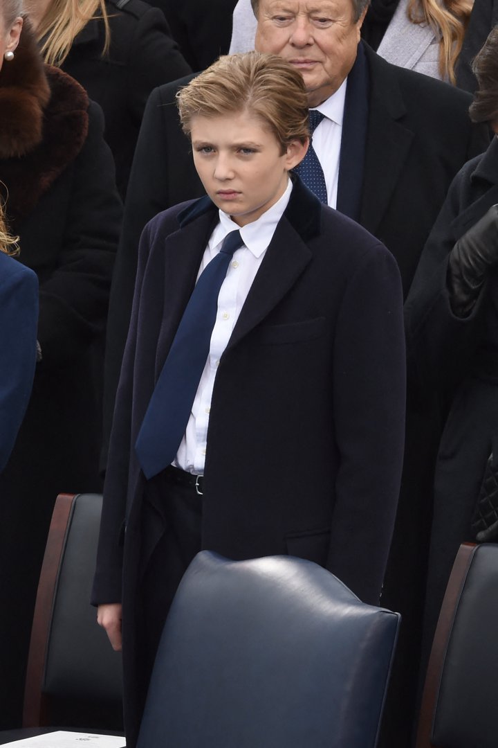 Barron Trump stands solemnly in a navy overcoat during the swearing-in ceremony at the U.S. Capitol. Dressed in a white shirt and blue tie, he appears focused amid the gathered crowd. The moment unfolds as Donald Trump is inaugurated as the 45th President of the United States.