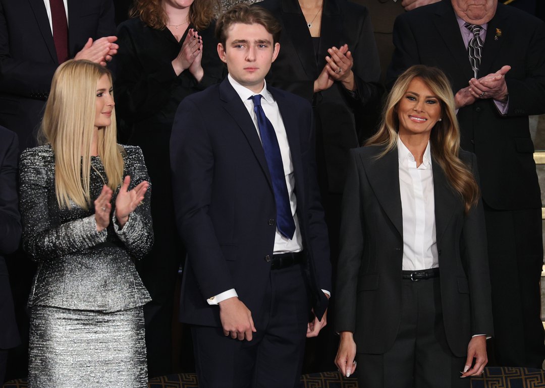 Ivanka Trump, Barron Trump, and First Lady Melania Trump stand during the State of the Union at the U.S. Capitol. Barron, in a navy suit and tie, looks ahead as applause fills the chamber around them. Melania smiles beside him while Ivanka turns toward the moment in the audience.