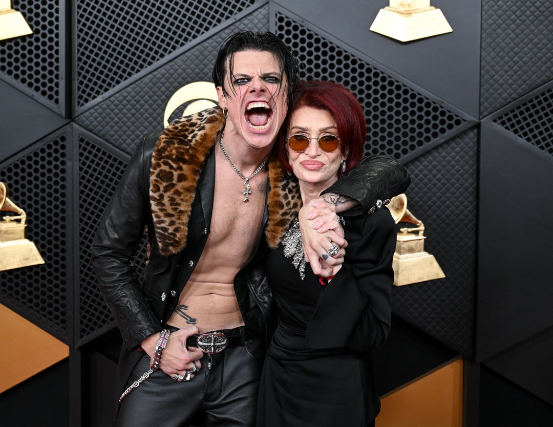 In another shot from the event, YUNGBLUD &mdash; joined by Sharon Osbourne &mdash; adjusts his stance, drawing attention to the full leather ensemble, including fitted black trousers and a statement belt buckle. The angle highlights the contrast between the sleek leather and the patterned collar, with his jewellery and makeup remaining focal points as he posed against the Grammys step-and-repeat.