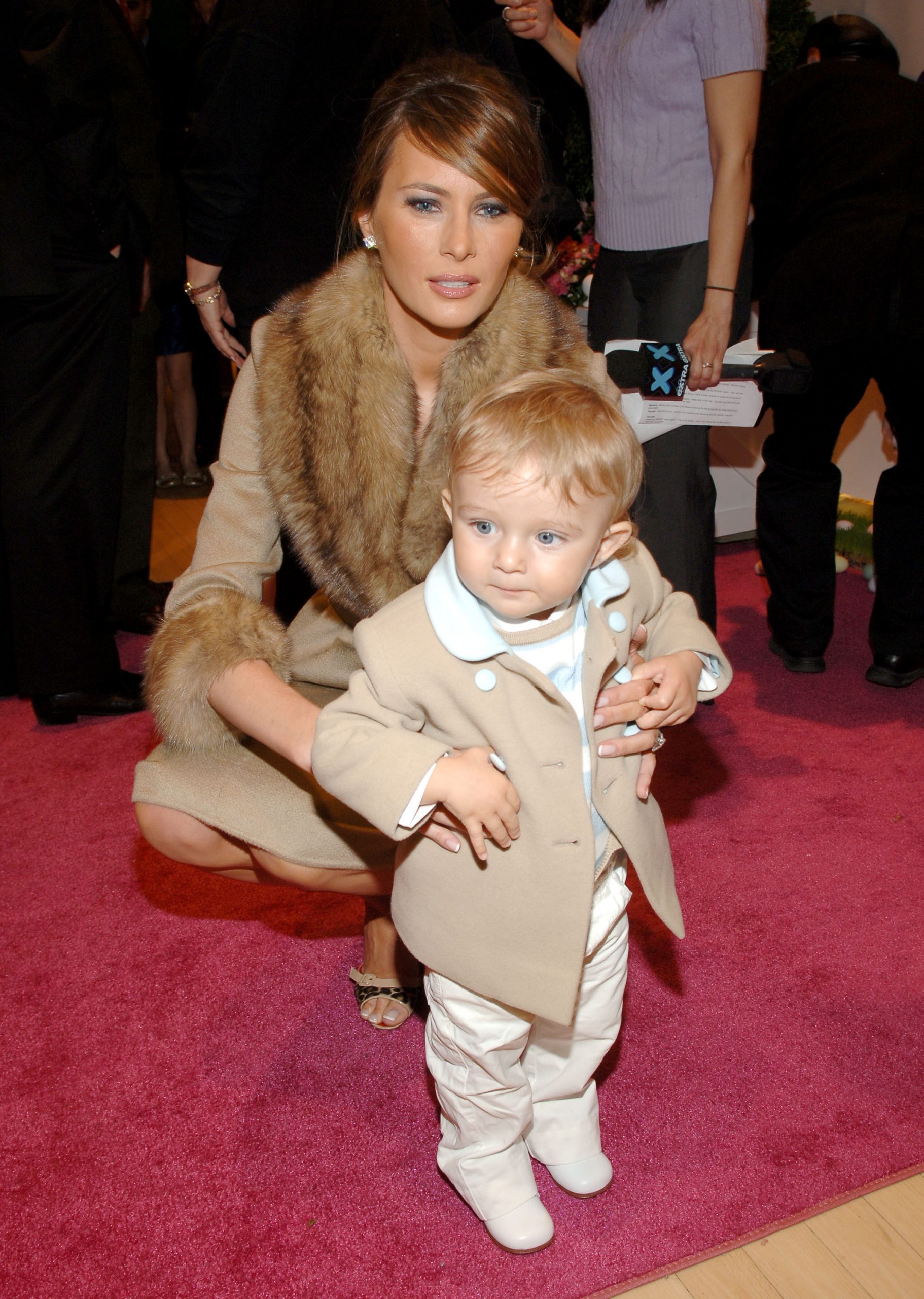 At FAO Schwarz in New York City, Melania Trump crouches beside baby Barron Trump on a pink carpet. Barron wears a tan coat with pale blue trim, while Melania pairs a fur-collared coat with open-toe heels.