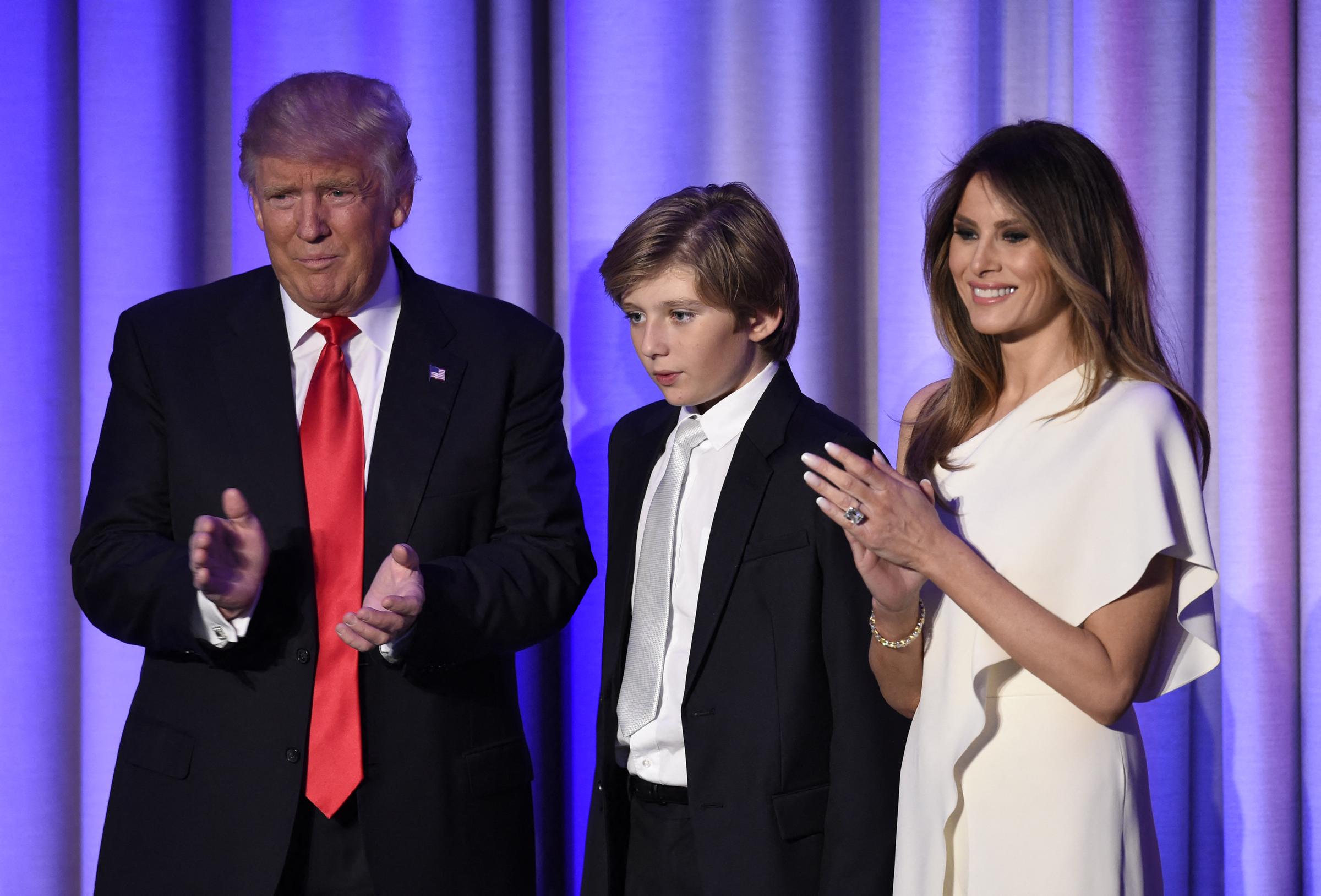 Donald Trump stands beside Melania Trump and their son, Barron Trump, at the New York Hilton Midtown on election night. Barron appears in a dark suit and tie as Melania, dressed in a white one-shoulder gown, applauds nearby.