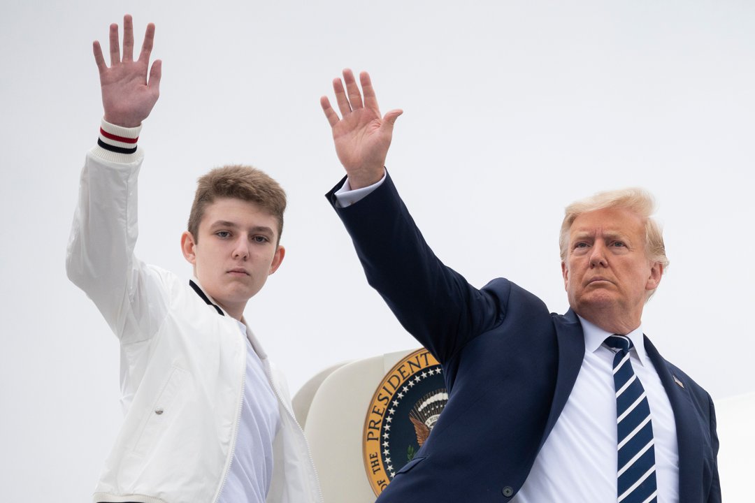 Donald Trump and his son Barron raise their hands in a wave while boarding Air Force One in Morristown, New Jersey. Barron stands beside his father in a white jacket, mirroring the gesture against an overcast sky. The presidential seal is visible behind them as they prepare for departure.
