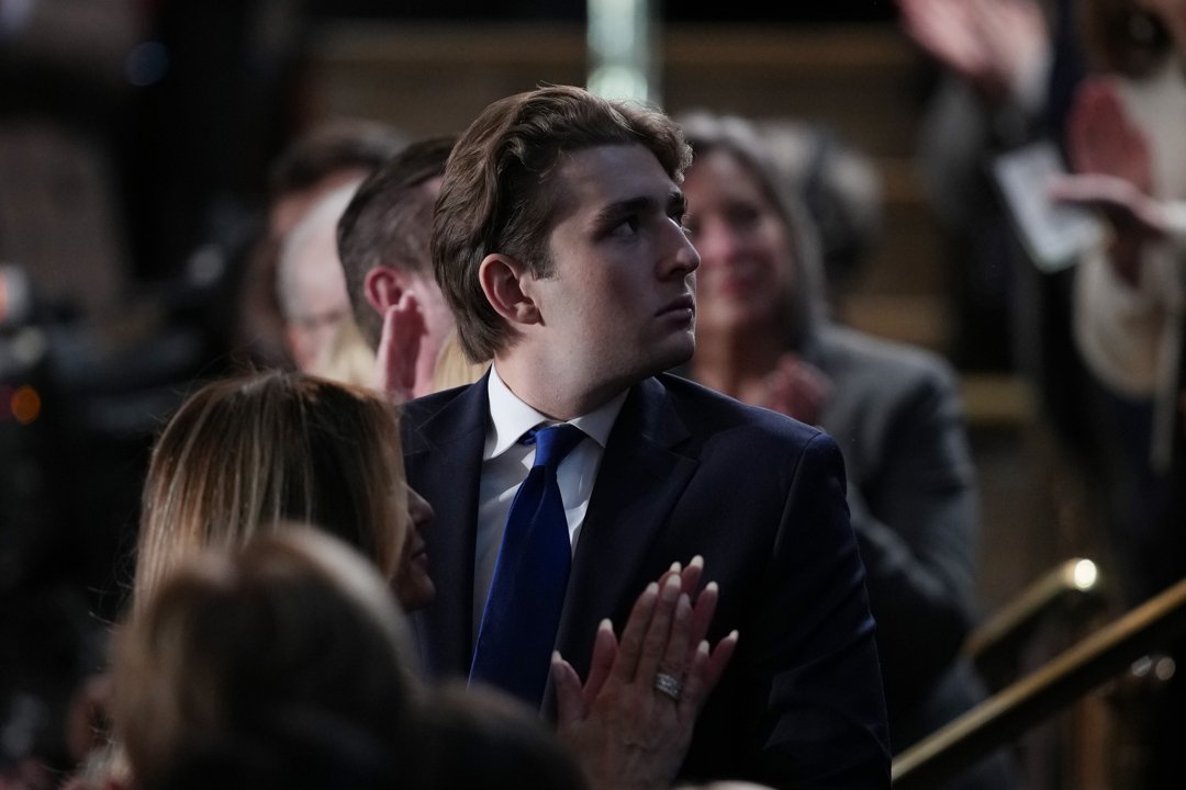 Barron Trump sits among attendees during the State of the Union address at the U.S. Capitol. Wearing a navy suit and blue tie, he turns his head as applause rises around him. Guests nearby clap while he looks toward the chamber floor.