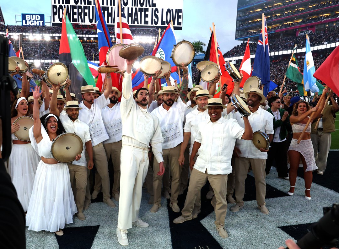 Bad Bunny performs with tambourine-holding dancers and flag bearers, raising his hand while surrounded by performers celebrating on the field during the halftime show.