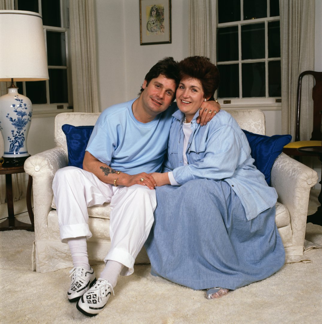Ozzy and Sharon Osbourne were photographed at home in the United States in 1987, seated closely on a cream sofa with their hands gently intertwined. Dressed casually in soft blues and whites, they leaned into one another with relaxed smiles, presenting a quieter portrait of life away from the stage. The domestic setting &mdash; framed artwork, lamplight, and evening windows &mdash; reflected a rare moment of stillness during their whirlwind years.