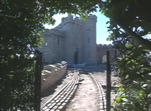 Framed by leafy branches, a narrow driveway curves towards the castle's entrance, drawing the eye to its battlements and central tower. The approach feels intentionally discreet, reinforcing the sense of retreat that has long defined Enya's life behind these walls. | Source: YouTube/AndreW