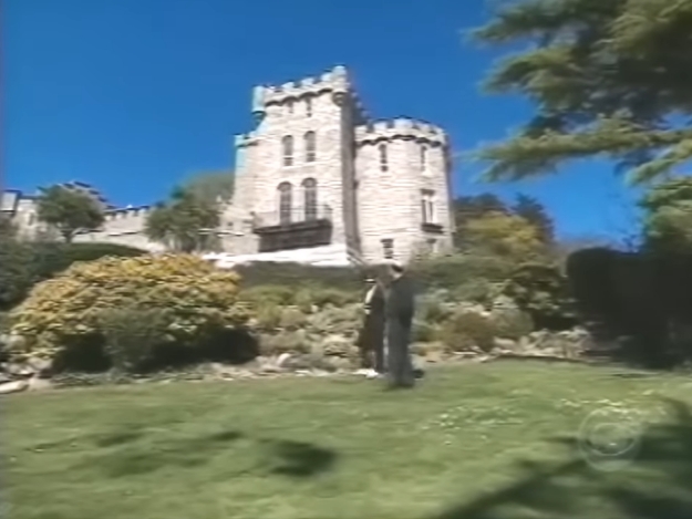 Set against a cloudless blue sky, the castle rises above manicured lawns and flowering shrubs, its turrets and arched windows evoking a romantic, almost storybook quality. The carefully tended grounds soften the fortress-like fa&ccedil;ade, revealing the balance between grandeur and domestic calm. | Source: YouTube/AndreW
