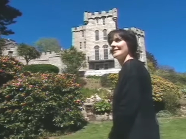 Captured mid-conversation, Enya smiles gently while positioned before vibrant shrubs and rolling lawns, the castle's crenellated roofline visible above her shoulder. The candid moment offers a rare glimpse of the singer outdoors at her coastal retreat, a place she has described as both refuge and inspiration. | Source: YouTube/AndreW