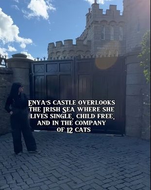 Standing before the tall, dark gates of her castle, a lone figure gazes up at the stone fa&ccedil;ade, dwarfed by its fortified walls and sunlit towers. The scene underscores the scale and privacy of the residence Enya made her own, a home designed as much for retreat as for creative reflection. | Source: YouTube/@beforethecoffin