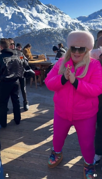 Gemma Collins claps her hands mid-dance while wearing oversized sunglasses, a fluffy pale pink hat, and matching pink leggings against a snowy mountain backdrop. | Source: Instagram/gemmacollins