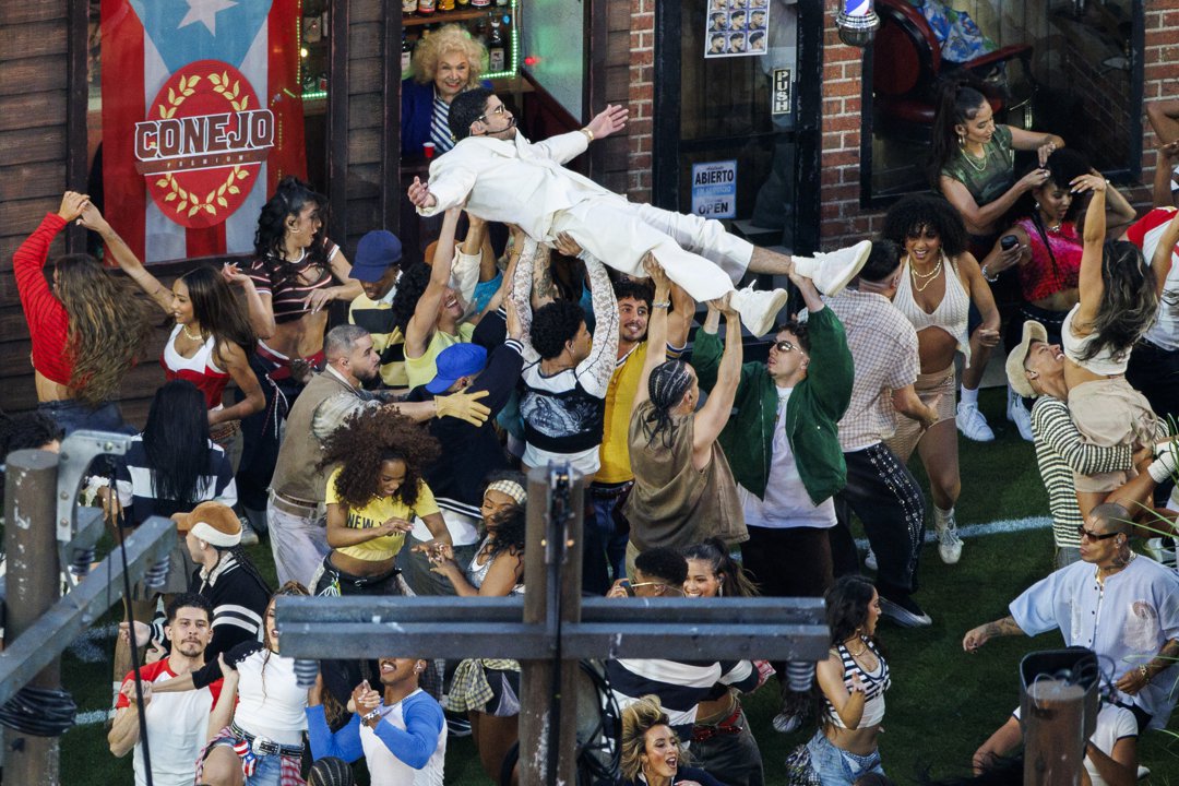 Bad Bunny is lifted by dancers while performing during the Super Bowl LX halftime show, surrounded by a crowd of performers in coordinated staging and movement.