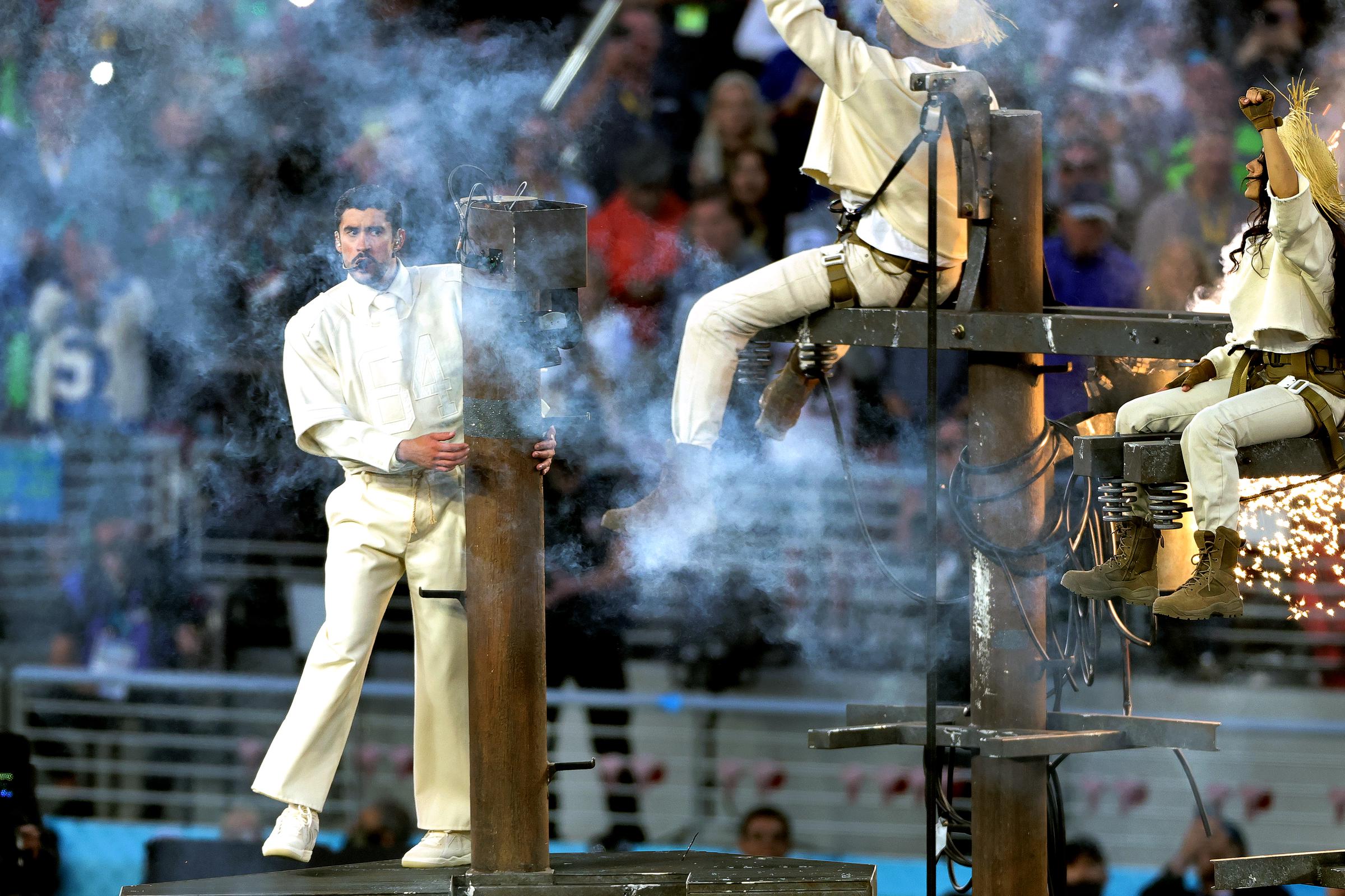 Bad Bunny appears amid smoke and stage effects, gripping a vertical prop as performers move around elevated structures during the halftime performance.