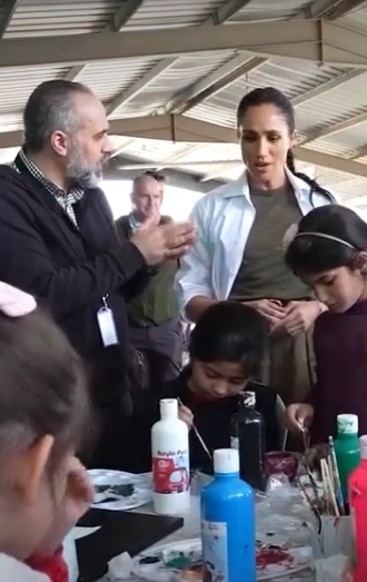 Meghan Markle leans in to observe children taking part in a hands-on activity during the couple's visit to Jordan, captured in a post dated February 26, 2026. Standing beside a local official who gestures mid-conversation, the Duchess watches closely as young participants concentrate on painting at a crowded table lined with brushes and bottles of colour. | Source: Instagram/pagesix