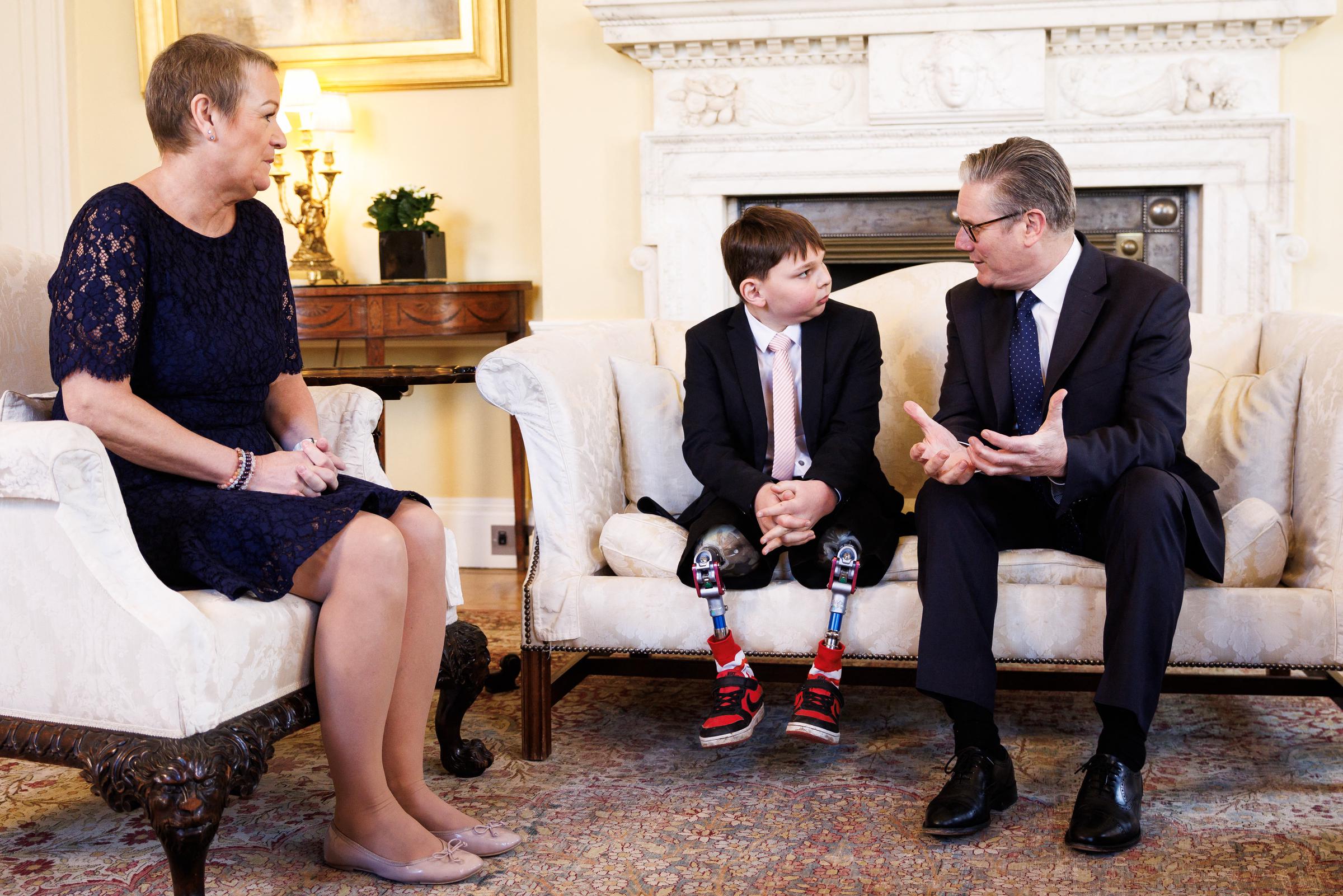 Starmer gestures as he speaks directly to Tony, who sits attentively beside him on a cream sofa, his prosthetic legs and red trainers visible beneath his smart black suit. Paula, in a navy lace dress, looks on from an armchair to the left, her hands clasped in her lap.