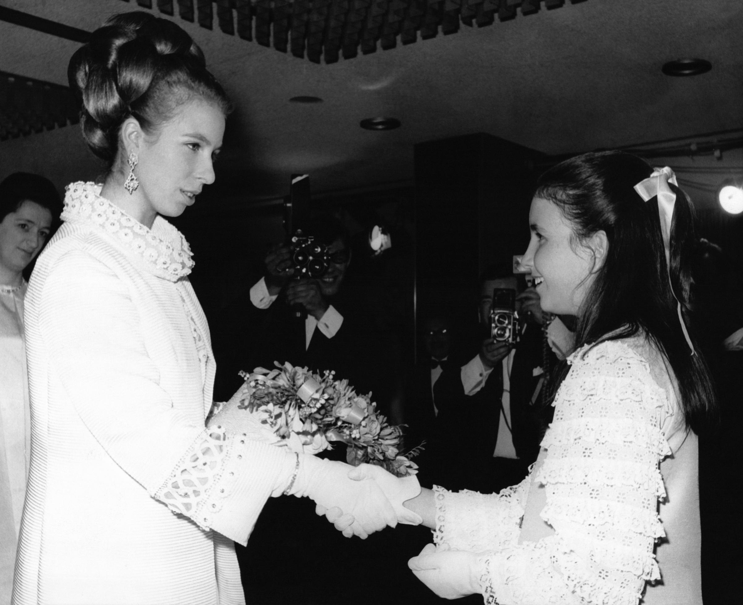 A beaming young girl presents an 18-year-old Princess Anne with a bouquet of flowers at the Run Wild, Run Free premiere at the Odeon Theatre in London in June 1969 — the very night the now-iconic cream coat made its debut. Anne, wearing white gloves, graciously accepts the flowers as photographers capture the moment behind them. The coat's ornate circular-patterned collar and matching geometric cuff detailing are clearly visible.