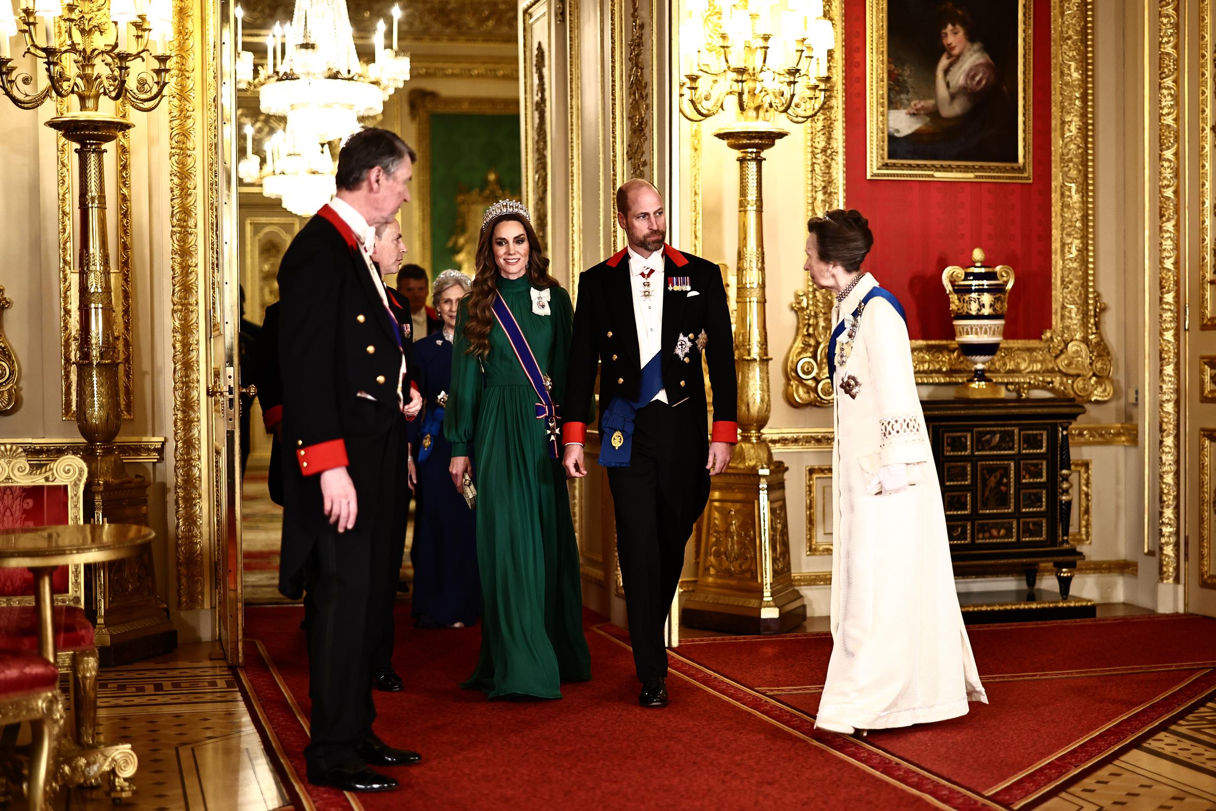 Princess Anne turns to greet the Prince and Princess of Wales as they enter St George's Hall, offering a rare view of the coat's back and the geometric cuff trim — while Catherine stuns in an emerald green gown and tiara alongside Prince William.